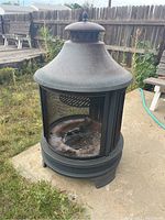 Full view of metal fire pit chiminea standing on concrete with lawn and wooden fence background. Shows cylindrical shape with chimney top and mesh door.