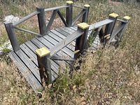 First large wooden garden bridge in yard with gold post caps, viewed angled from side and top showing construction and condition.