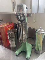 Milkshake maker with chrome head and green base, three stainless steel cups stacked beside it, clear straw holder with colorful plastic straws, and Betty Crocker cookbooks in background.