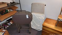 Three beige fabric-covered TV trays on a brown metal rack with wheels, next to two white plastic folding tables and a wooden box.