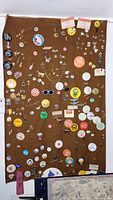 Full view of brown fabric board with a large assortment of vintage pins and buttons attached, showing a variety of colors and designs including political buttons and character pins.