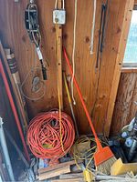 Photo shows a wooden shed wall with a metal hand winch hanging, several pulleys attached, long yellow and red wooden poles, a broom with orange head, some coiled orange extension cords, and a wooden stack on the floor.