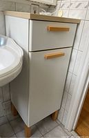 Side view of white bathroom cabinet with light wood top, drawer and door, wooden handles, positioned beside a sink