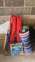 Two red camping chair bags, two rolled ground mats, two umbrellas with clamps, two green pillows, and a boxed pool solar cover visible on floor against wall.
