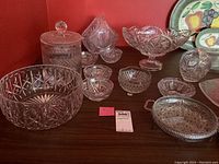 Image showing the twelve pieces of assorted clear crystal serving ware including biscuit jar, bowls, and nappies on a wooden surface with patterned plates in background.