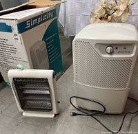Photo showing both the Simplicity dehumidifier with its original box and the Airworks electric heater side by side on the floor.