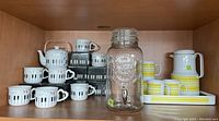 Shelf with ceramic keyboard-themed teapot and mugs, plastic beverage carafe with matching yellow/white mugs and tray, and a glass beverage dispenser