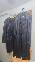 Two black leather trench coats hanging side by side. One shorter with hood and drawstring waist, the other longer with button closure.