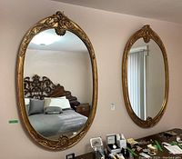 Front view of two large oval wall mirrors with ornate gold-painted carved frames hanging on a wall.