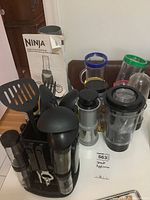 Various Ninja and Nutri Ninja blending containers with different colored lids, black utensil caddy holding kitchen utensils like spatula, ladle, and peeler, displayed on wooden surface with Ninja system box in background.
