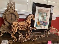 Photo showing wooden mechanical clock with detailed gears and scrollwork beside mechanical horse and several small mechanical figures and parts on a wooden surface.