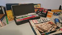 Photo showing Mah-Jongg set in black case, two wooden cribbage boards, and Upwords, Eclipse, Yum, and Trivial Pursuit boxed games in background with chess set on left