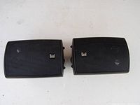 Photo showing two black compact Dual indoor/outdoor speakers placed side by side. The speakers have rounded edges and a front grill showing the Dual logo.