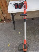 Photo showing the orange and black Craftsman cordless grass trimmer leaning against a wall next to a white folding table. On the table are two black rechargeable batteries and two battery chargers with cords.