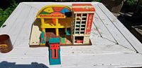 Color photo showing Fisher Price toy garage playset on white wood table outdoors. The set has a yellow spinning dial, a white and red tower labeled "FLEX-TO-LIFT" with a working elevator hoist, green ramp, and multiple parking areas.