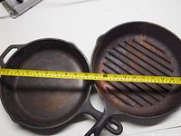 Two Lodge cast iron skillets side by side on a white surface with measuring tape showing approximately 10 inches diameter.