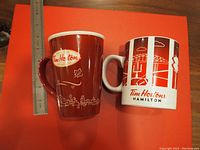 Both Tim Hortons Hamilton mugs side by side on a red surface with a ruler to show scale, highlighting exterior designs and colors.
