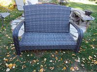 Full front view of a dark brown wicker/rattan patio loveseat sofa placed on a stone patio with fallen leaves around.