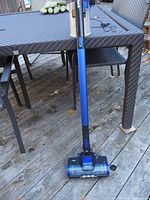 Full view of the Shark Pet Plus cordless vacuum standing upright on a wooden deck with an outdoor table and chairs in the background.