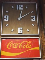 Photo showing vintage Coca-Cola analog square wall clock with raised white numbers and white hour markers on brown background. Below is a rectangular Coca-Cola sign with red background and yellow logo.