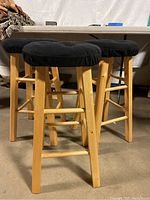 Three wooden bar stools with round black fabric seats shown from side angle against a background of a folding table and other items in storage.