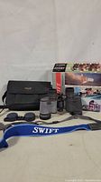 Swift binoculars with textured gray body, black eyepieces, blue neck strap, lens caps, and carrying case alongside original box.