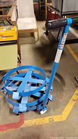 Blue Clamp & Go Drum Dolly with circular clamp and one handle with black grip, placed on warehouse floor.
