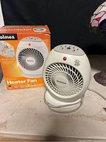 Holmes compact heater fan in off-white and gray placed next to its original orange and black box with product image and branding.