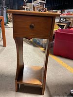 Front view of the small wooden night stand showing a single drawer with a round knob and a lower open shelf. The wood has a natural finish with a smooth surface and visible grain.