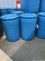 Two blue plastic barrels standing upright on an outdoor pavement area, grouped with similar barrels in the background.