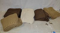 Five folded knitted blankets in shades of brown, beige, and cream arranged side by side on the floor.
