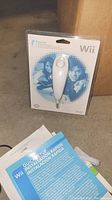 White Nintendo Wii Nunchuk controller in original sealed packaging on concrete floor, with Wii Quick Start Guides partially visible in front.