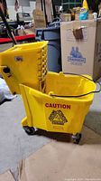 Yellow plastic mop bucket labeled with caution warning, fitted with caster wheels for portability.