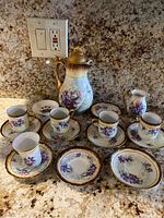 Chocolate set displayed on granite countertop including tall chocolate pot, creamer, 5 cups with saucers, and 2 small bowls, decorated with purple and gold floral motif.