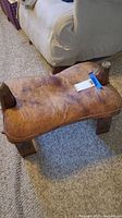 Angled side view of leather camel saddle stool with wooden legs on carpeted floor, showing wear and general shape.