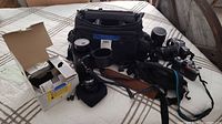 Lot contents on bed including black camera bag, three cameras with attached lenses, various lenses with caps and straps, and cardboard box.