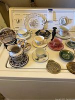 Full view of assorted cups, saucers, decorative plate, and jug arranged on a white appliance surface showing various colors and patterns.
