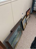 Side view showing five framed paintings on the floor leaning against the wall, varying in size and frame style.