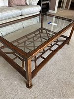 Glass top coffee table with visible carved wooden lattice base beneath glass, medium brown wood with natural wear, photographed in living room setting.