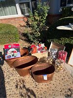Photo showing three brown metal buckets, assorted Christmas ornaments in plastic containers, a box with ornaments, and a boxed inflatable Christmas camper decoration on a textured concrete surface outdoors.
