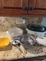 Aroma crockpot with green ivy design and glass lid, CorningWare casserole dish with floral design and glass lid, stainless steel colander, small yellow plastic colander or strainer, and crockpot lid knob.