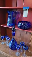Full view of cobalt blue pitcher, bowl, decanter, and six goblets on wooden shelves.