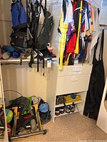 Photo showing blue and gray backpacks, colorful biking jerseys hanging, bike shoes on shelves, and black ski bib pants hanging.