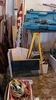 Assorted wood pieces, curtain rods, folded American flag, extension cords, and blue case for Nokita saw. Items arranged near wall and on floor.