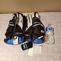Pair of black Bauer Men's Challenger SR hockey gloves with blue inner lining, displayed flat next to a water bottle for size reference. Gloves have tags attached.