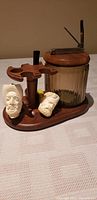 Full shot showing wooden pipe stand with two white hand-carved Meerschaum style pipes with bearded faces, a dark briar pipe, and glass humidor with wooden lid and metal clasp