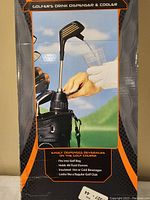 Close-up photo of the product showing dispensing spout shaped like a golf club head pouring beverage into a cup. Packaging text highlights product features such as fitting into a golf bag, holding 48 fluid ounces, being insulated, and resembling a regular golf club.
