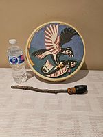 Front view of the 12" Indigenous hand drum with colorful Haida eagle design and handmade beater placed in front for scale with water bottle.