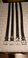 Four black men's tooled leather belts laid out full length on table, showing metal buckles and length variation.
