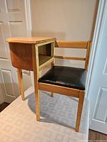 Side angled view of the wooden telephone table with attached chair showing the storage compartment and the black padded seat.
